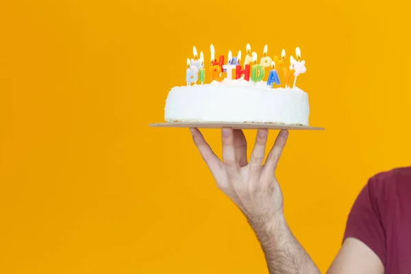 Hands holding a birthday cake with candles and the inscription birthday on a yellow background. Congratulations on the anniversary and birthday. Copyspace.