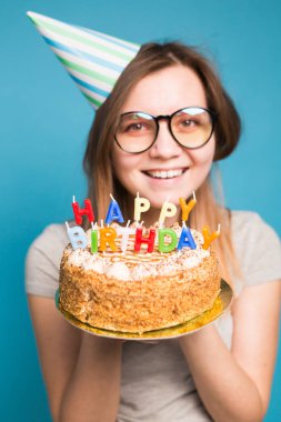 Mavi arka planda büyük bir Doğum günü pastası tutan bir kağıt şapka ve gözlük çılgın komik kız. Şaka ve selamlar kavramı.