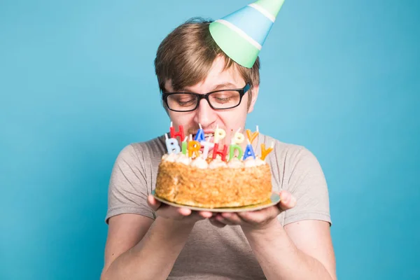 Loco joven divertido en gafas y papel sombreros de felicitación celebración de pasteles feliz cumpleaños de pie sobre un fondo azul . —  Fotos de Stock