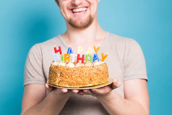 Divertente giovane studente pazzo in un cappello di saluto gioisce accanto alla torta con l'iscrizione buon compleanno in piedi su uno sfondo blu. Concetto di saluto. Copia spazio — Foto Stock