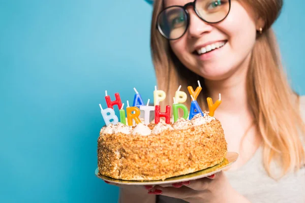 Fille drôle folle dans un chapeau en papier et des lunettes tenant un gros gâteau d'anniversaire sur le fond bleu. Concept de farce et salutations . — Photo