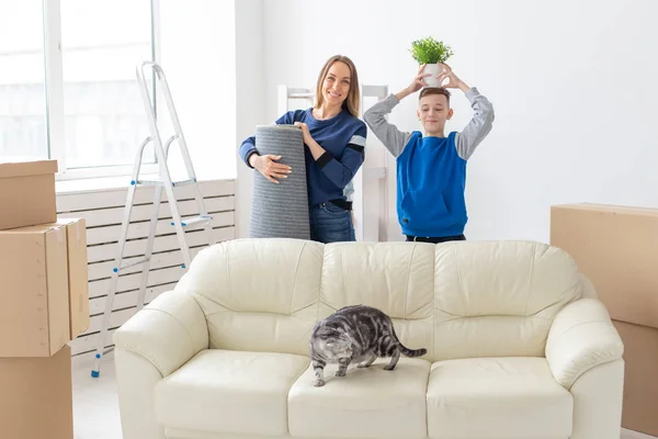 Молодые симпатичные мать и сын-одиночка рады переезду в новый дом, держа в руках одноухий шотландский кот и кастрюлю зелени. Концепция новоселья и расширения семейного пространства . — стоковое фото