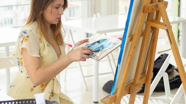Art school, creativity and leisure concept - student girl or young woman artist with easel, palette and paint brush painting picture at studio, close-up