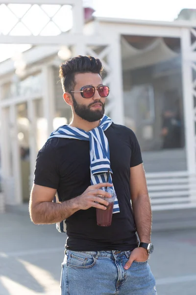 Retrato de um jovem árabe alegre positivo com um copo de suco com uma palha enquanto caminha no parque em um dia de verão ensolarado quente. O conceito de descanso após estudo e trabalho nos fins de semana . — Fotografia de Stock