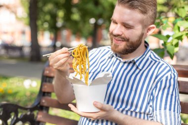 Güneşli bir yaz gününde bir park bankında otururken bir öğle yemeği kutusundan Çin erişte yiyen genç yakışıklı hipster adam Yan görünümü. Sağlıklı ve besleyici bir atıştırmalık kavramı.