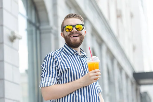 Retrato de un joven hipster borroso y elegante con barba y bigote sosteniendo jugo de naranja fresco en sus manos. Concepto de snack saludable . — Foto de Stock