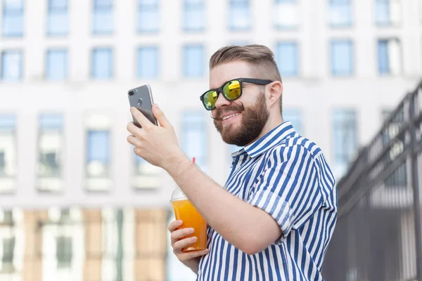 Vista lateral de un joven y elegante hombre hipster con gafas y un bigote y jugo en sus manos charlando en las redes sociales usando un teléfono inteligente e internet inalámbrico en un cálido día de verano . — Foto de Stock