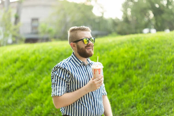 Vista lateral de un joven elegante con una barba sosteniendo un batido y admirando las vistas de la ciudad caminando en el parque en un cálido día de verano. El concepto de descanso y relajación . — Foto de Stock