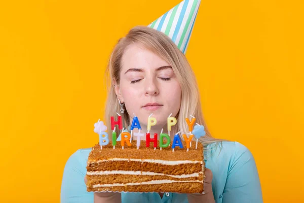 Retrato de close-up de uma garota positiva engraçada com um boné de papel segurando um bolo de parabéns em suas mãos em um fundo amarelo. Conceito e diversão e celebração. Espaço publicitário . — Fotografia de Stock