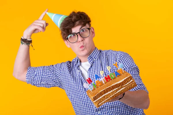 Retrato de un tipo positivo divertido con una gorra de papel y vasos sosteniendo un pastel de felicitación en sus manos sobre un fondo amarillo. Concepto y diversión y celebración. Espacio publicitario . — Foto de Stock