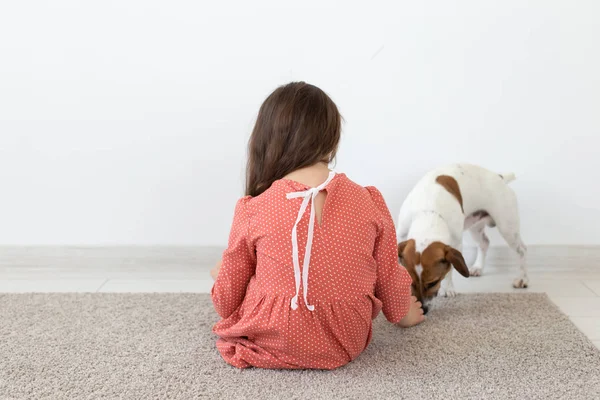 Vista trasera de una niña pequeña con un vestido de lunares rojos sentada en el suelo junto a su amado perro Jack Russell Terrier sobre un fondo blanco. El concepto de productos para niños . — Foto de Stock