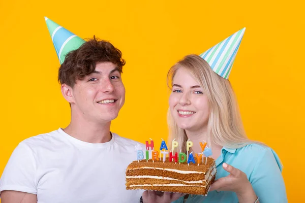 Funny Nerd mężczyzna i kobieta noszą wakacje czapki i okulary gospodarstwa tort urodzinowy ze świecami na żółtym tle — Zdjęcie stockowe