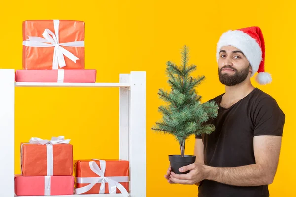 Jonge positieve man met een baard en kerstman hoed staat in de buurt van stand met geschenken en kerstboom op gele achtergrond. Concept van felicatie geschenk en kortingen voor Kerstmis en Nieuwjaar. — Stockfoto