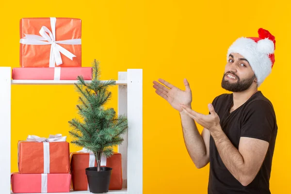 Jeune homme positif avec une barbe et le chapeau du Père Noël est debout près du stand avec des cadeaux et un arbre de Noël sur fond jaune. Concept de cadeau de félicitations et de réductions pour Noël et le Nouvel An . — Photo