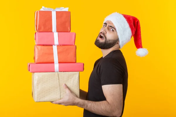 Niezadowolony młody człowiek z brodą w kapeluszu Świętego Mikołaja posiada pięć pudełek upominkowych stwarzających na żółtym tle. Koncepcja prezentów i pozdrowienia dla świąt Bożego Narodzenia i nowego roku. — Zdjęcie stockowe
