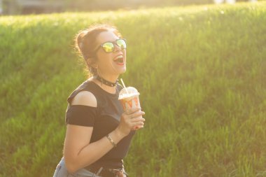 Şık punk giysileri ve gözlük sıcak bir yaz akşamı parkta bir yürüyüş sırasında elinde bir milkshake tutan olumlu neşeli genç kadın. Şehirde rahatlama ve eğlence kavramı.