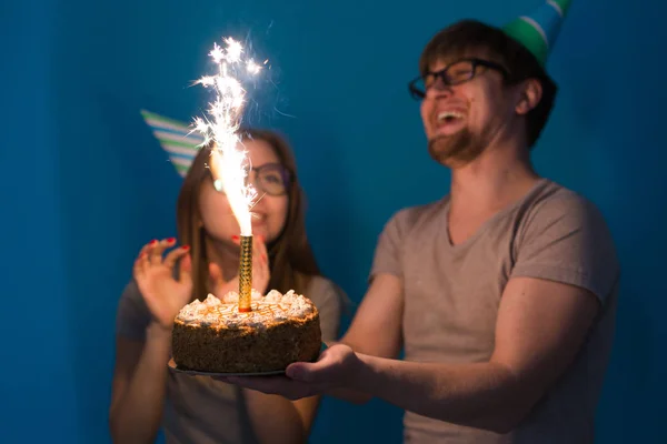 紙の帽子とケーキで面白い若いカップルは愚かな顔を作り、青い背景に立っている間、幸せな誕生日を願っています。おめでとうとだまさの概念. — ストック写真