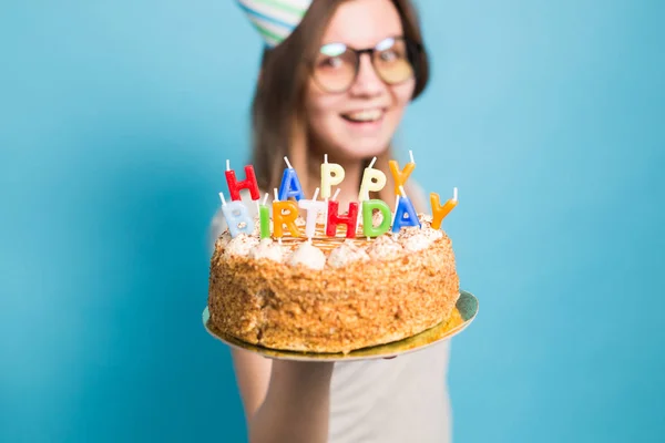 Γοητευτικό χαρούμενο τρελό νεαρό κορίτσι μαθητής σε συγχαρητήριο καπέλο χαρτί κρατώντας μια χαρούμενη τούρτα γενεθλίων στα χέρια της στέκεται σε ένα μπλε φόντο. Διαφημιστικός χώρος. — Φωτογραφία Αρχείου