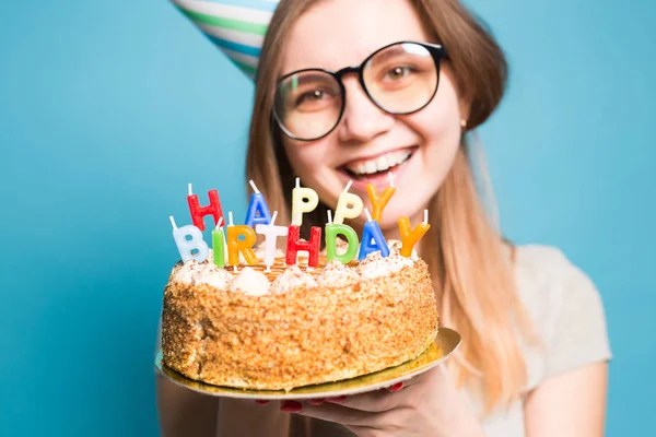 Közelről vicces pozitív lány a szemüveg és üdvözlő papír kalap gazdaság boldog születésnapi torta a kezében állt a kék háttér. — Stock Fotó