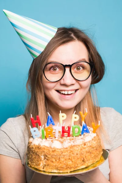 Bájos vidám őrült fiatal lány diák gratuláló papír kalap gazdaság boldog születésnapi torta a kezében állva ellen kék háttérben. Reklámfelület. — Stock Fotó