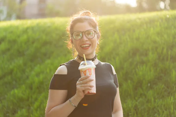 Portret pozytywnej młodej dziewczyny w punkowych ubraniach i okularach z Milkshake w rękach stwarzających w letnim parku w ciepłym letnim wieczorem. Koncepcja urlopów. — Zdjęcie stockowe
