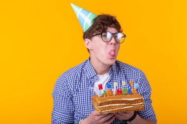 Kağıt kutlama şapka deli neşeli genç adam sarı bir arka plan üzerinde duran kek mutlu doğum günü tutan. Jubilee tebrikler kavramı.