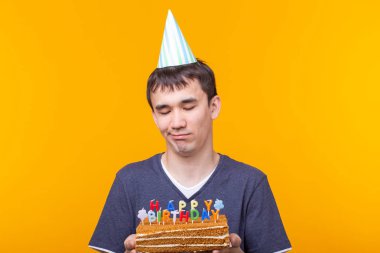 Bir şapka ve yanan bir mum ve elinde bir ev yapımı pasta sarı bir arka plan üzerinde poz ile Pozitif komik genç Asya adam. Yıldönümü ve doğum günü konsepti.
