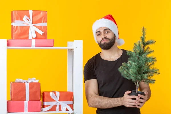 Jonge positieve man met een baard en kerstman hoed staat in de buurt van stand met geschenken en kerstboom op gele achtergrond. Concept van felicatie geschenk en kortingen voor Kerstmis en Nieuwjaar. — Stockfoto