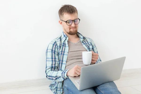 Pozytywny młody człowiek w casual ubrania surfowanie po Internecie w poszukiwaniu nowych mieszkań siedzi na podłodze w pustym pomieszczeniu. Koncepcja znalezienia mieszkania za pomocą Internetu i laptopa. — Zdjęcie stockowe