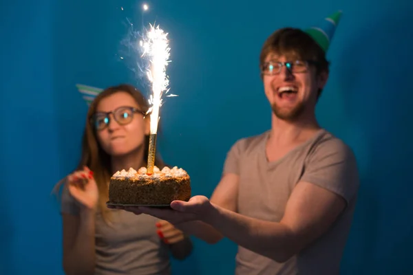 Alegre jovem casal encantador cara e bonito menina em chapéus de papel fazer cara tola e segure em suas mãos um bolo de aniversário em pé sobre um fundo azul . — Fotografia de Stock