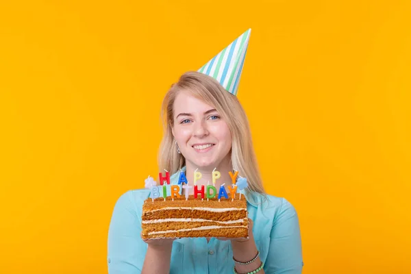 Gek vrolijke jonge vrouw in papier felicitatie hoed houden taarten gelukkige verjaardag staande op een gele achtergrond. Jubileum Gefeliciteerd concept. — Stockfoto