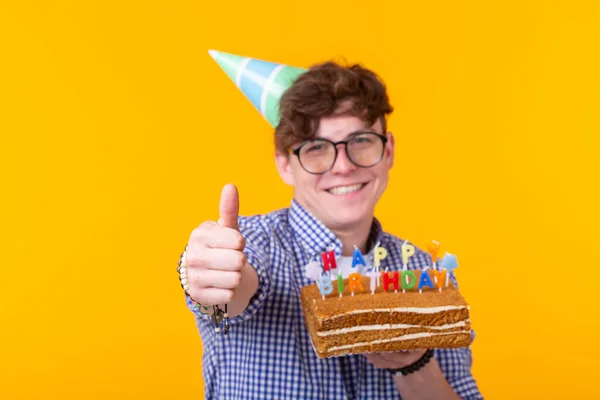 Retrato de um cara positivo engraçado com um boné de papel e óculos segurando um bolo de parabéns em suas mãos em um fundo amarelo. Conceito e diversão e celebração. Espaço publicitário . — Fotografia de Stock