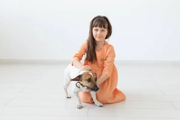 Pequeña chica encantadora con el pelo oscuro en un vestido largo está sentado al lado del perro Jack Russell Terrier en el suelo de su habitación. El concepto de inocencia infantil y cuidado infantil. Espacio de copia . — Foto de Stock