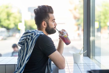Yakışıklı genç hipster adam bir kafede kokteyl içme ve pencereye bakarak.