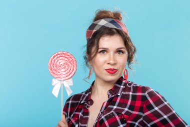 Elinde renkli lolipop tutan ve mavi arka planda poz birini yalayan retro giysiler içinde Büyüleyici genç kadın. Tatlılar ve tatiller için aşk kavramı.