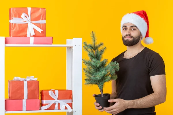 Jonge positieve man met een baard en kerstman hoed staat in de buurt van stand met geschenken en kerstboom op gele achtergrond. Concept van felicatie geschenk en kortingen voor Kerstmis en Nieuwjaar. — Stockfoto