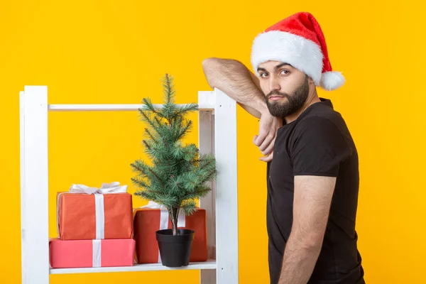 Jonge positieve man met een baard en kerstman hoed staat in de buurt van stand met geschenken en kerstboom op gele achtergrond. Concept van felicatie geschenk en kortingen voor Kerstmis en Nieuwjaar. — Stockfoto