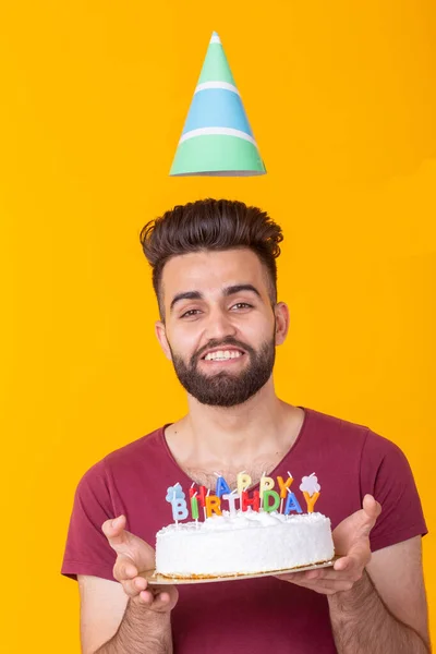 Embaraçado jovem hipster masculino com barba segurando um bolo de aniversário em suas mãos e olhando atenciosamente para ele posando em um fundo amarelo. Conceito de tempo voa rapidamente por . — Fotografia de Stock