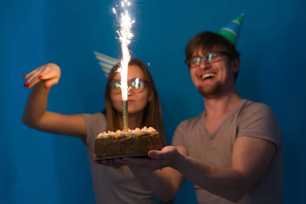 Jóvenes estudiantes alegres encantadora chica y buen chico en gorras de papel de saludo sosteniendo un pastel con una vela chispas de bengala. Concepto de felicitación por el cumpleaños y aniversario —  Fotos de Stock