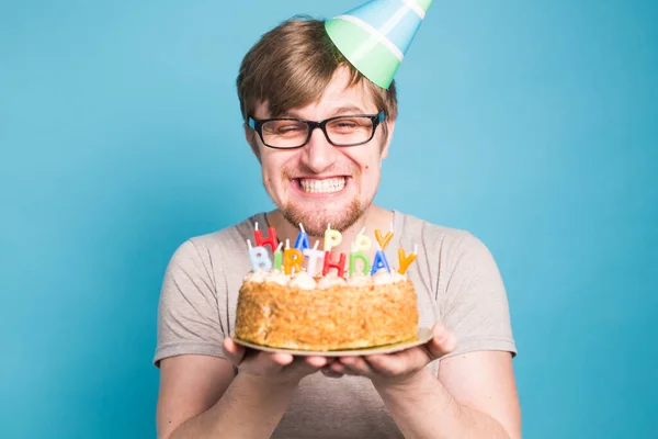 Estudante jovem louco engraçado em um chapéu de saudação se alegra ao lado do bolo com a inscrição feliz aniversário de pé em um fundo azul. Conceito de saudações. Espaço de cópia — Fotografia de Stock