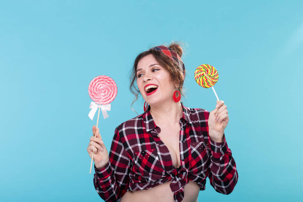 Charming young woman in retro clothes holding colorful lollipops in her hands and licking one posing on blue background. Concept of love for sweets and holidays.