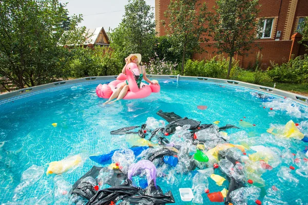 Problema de lixo, reciclagem de plástico, poluição e conceito ambiental - mulher boba nada e se divertir em uma piscina poluída. Garrafas e sacos de plástico flutuam perto dela — Fotografia de Stock