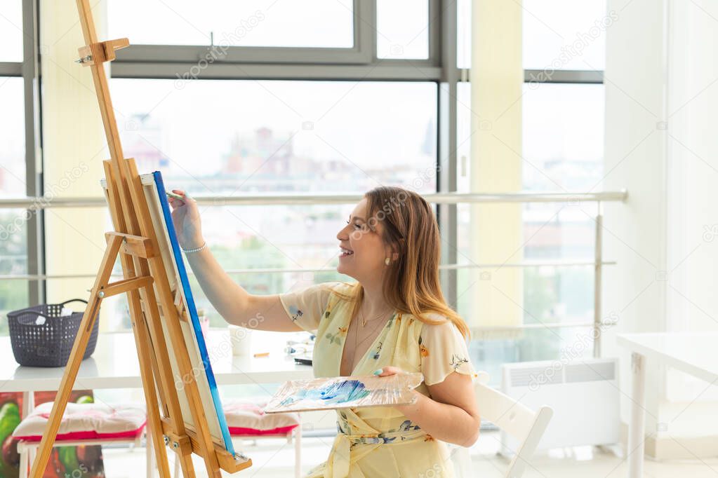 Art school, creativity and leisure concept - student girl or young woman artist with easel, palette and paint brush painting picture at studio