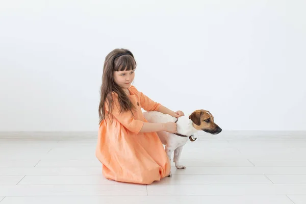 Liten charmig flicka med mörkt hår i en lång klänning sitter bredvid hunden Jack Russell Terrier på golvet i hennes rum. Begreppet barn oskuld och barnomsorg. Copyspace. — Stockfoto