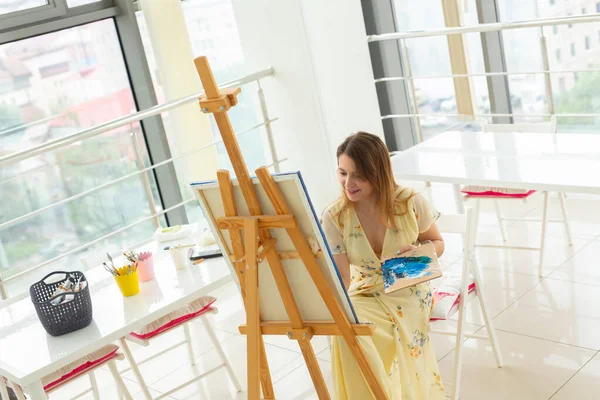 Art class, drawing and creativity concept - Female student sitting in front of easel with palette and paintbrush — Stock Photo, Image