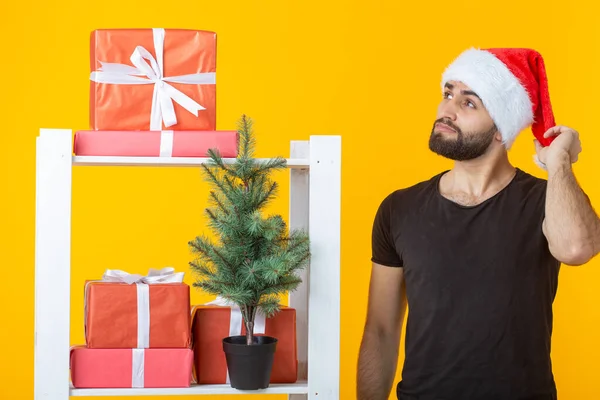 Jonge positieve man met een baard en kerstman hoed staat in de buurt van stand met geschenken en kerstboom op gele achtergrond. Concept van felicatie geschenk en kortingen voor Kerstmis en Nieuwjaar. — Stockfoto