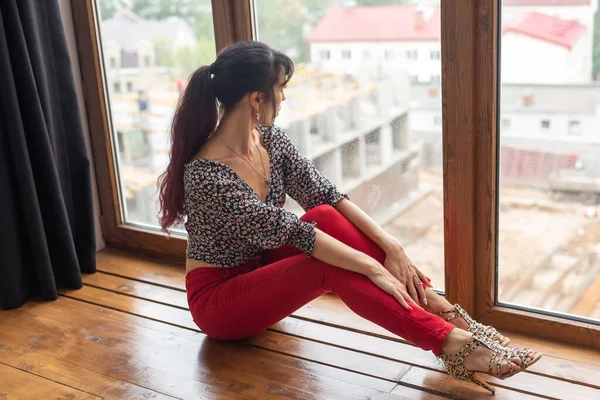 Tagträumerei, Nachdenklichkeit und Einsamkeit - junge kaukasische Frau sitzt in ihrem Wohnzimmer und blickt durch ein großes Fenster hinaus — Stockfoto