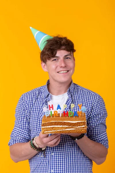 Pozitív vicces fiatal fickó egy sapkát és egy házi készítésű sütemény a kezében pózol egy sárga háttér. Évforduló-és születésnapi koncepció. — Stock Fotó
