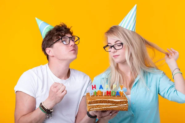 Joven pareja alegre chico encantador y linda chica en sombreros de papel hacen cara tonta y tienen en sus manos un pastel con la inscripción cumpleaños de pie sobre un fondo amarillo. Concepto saludos y —  Fotos de Stock