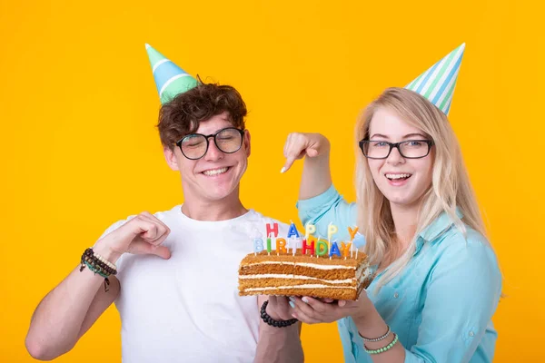 Веселая молодая пара очаровательный парень и милая девушка в бумажных шляпах делают глупое лицо и держать в руках торт с надписью день рождения стоя на желтом фоне. Концепция приветствия и — стоковое фото